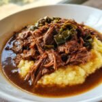 Shredded Instant Pot Mississippi Pot Roast served over creamy yellow grits with rich gravy and pepperoncini slices.