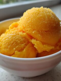 Close-up of bright orange, icy Mango Sorbet scooped into a small white bowl.