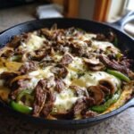 Close-up of a sizzling Philly Cheesesteak Skillet topped with melted white cheese, steak strips, green peppers, and mushrooms.