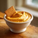A small white bowl filled with bright orange Pumpkin Peanut Butter Dip, featuring a graham cracker dipped inside.