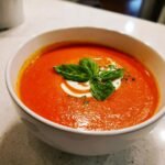 A close-up of vibrant orange Roasted Tomato Soup garnished with fresh basil leaves and a swirl of cream.