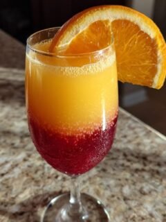 A layered Sunrise Mimosa cocktail showing red, orange, and yellow layers, garnished with an orange slice.