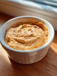 A small white ramekin filled with creamy, swirled Peanut Butter Dip, sitting on a sunlit wooden surface.