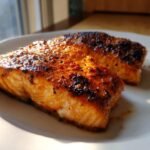 Two beautifully seasoned and caramelized fillets of Air Fryer Salmon resting on a white plate, highlighted by natural sunlight.