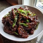 A white bowl filled with sticky, dark glazed Mongolian Beef, garnished generously with sliced green onions and sesame seeds.