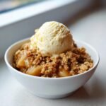 A close-up of a warm serving of Apple Crumble topped with a scoop of melting vanilla ice cream in a white bowl.