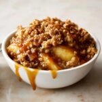 Close-up of a serving of rich Caramel Apple Crisp topped with gooey caramel sauce dripping down the side.