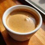 Close-up of a small white cup filled with espresso showing rich, speckled brown crema, sitting on a wooden surface.