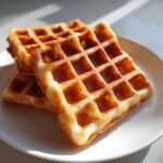 Close-up of two golden brown Banana Waffles stacked on a white plate, beautifully lit by sunlight.