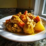 A close-up of rich, yellow Jamaican Curry Chicken served with chunks of carrot and potato on a white plate.