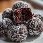 Close-up of several moist Brownie Truffles rolled in white powdered sugar, one is broken open showing the dark interior.