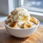 A white bowl filled with Patti Labelle Banana Pudding topped with a scoop of melting vanilla ice cream and caramelized banana slices.
