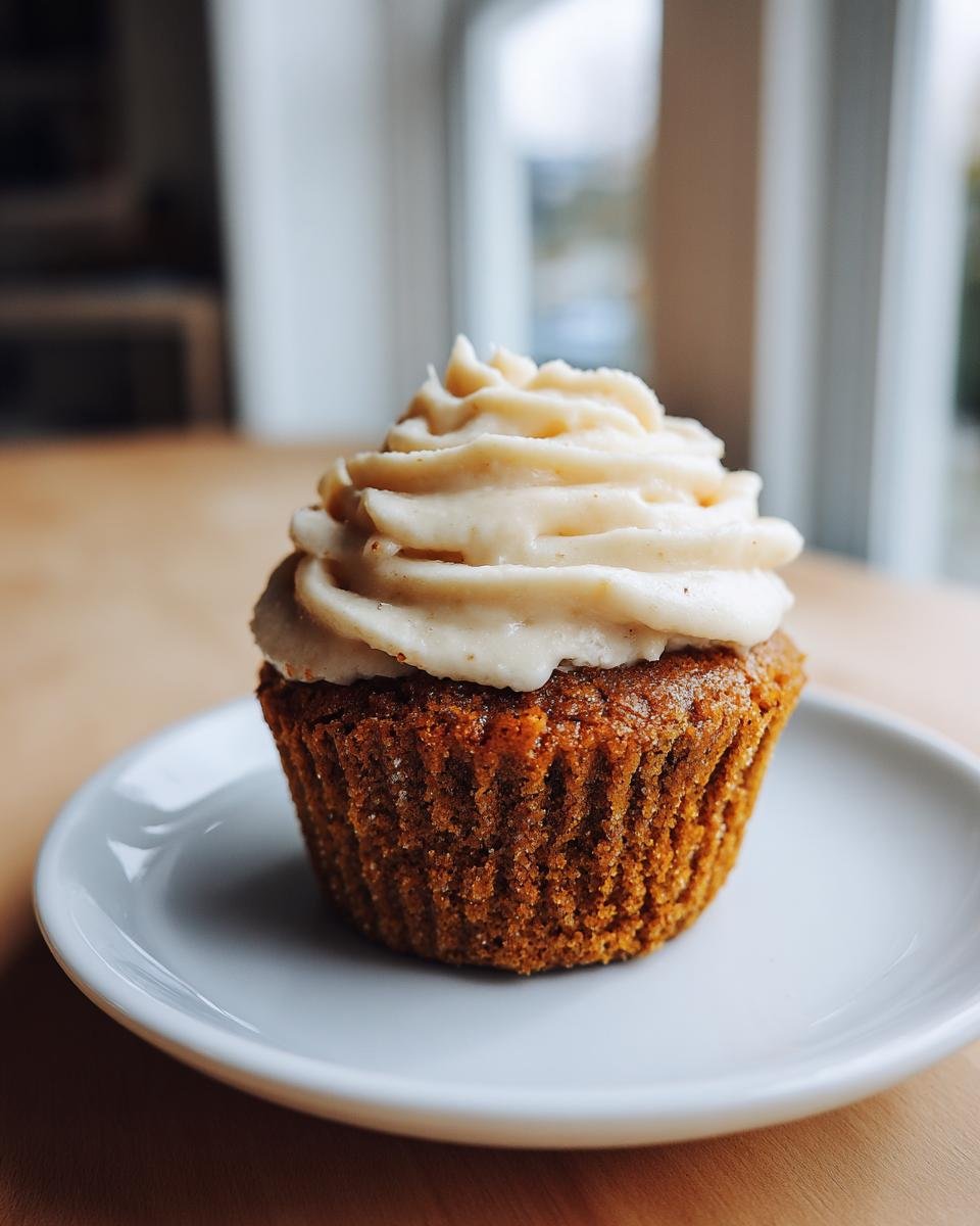 A single, perfectly baked Pumpkin Cupcake With Maple Frosting sitting on a small white plate.