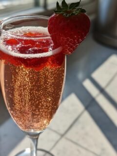 A flute glass filled with a sparkling, pink Rossini Cocktail, garnished with a fresh strawberry.