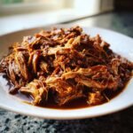 A close-up of tender, shredded Braised Pork Shoulder coated in rich, dark sauce, served on a white plate.