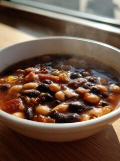 A steaming white bowl filled with a rich, dark stew containing hominy and black beans, set on a wooden surface near a window.