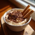 A white bowl filled with creamy Tiramisu Dip, topped with chocolate shavings and three ladyfinger cookies for dipping.