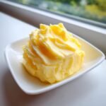 A swirl of bright yellow, whipped Honey Butter served in a small white dish near a window.
