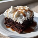 A close-up of a moist slice of Baileys Chocolate Poke Cake topped with whipped cream and caramel drizzle.