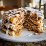 A tall stack of fluffy Cinnamon Roll Pancakes, cut open to show cinnamon filling, drizzled heavily with white icing.