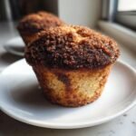 A close-up photo of a freshly baked Cinnamon Streusel Muffin with a thick, dark brown crumb topping on a white plate.
