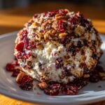 A close-up of a festive Cranberry Pecan Cheese Ball coated in dried cranberries and chopped pecans on a white plate.