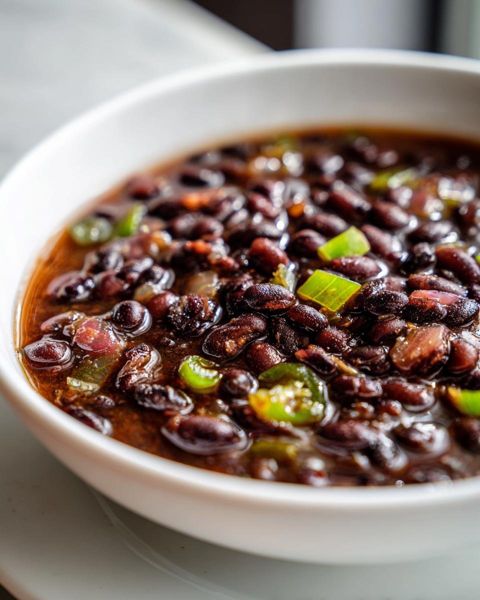 A close-up, macro shot of savory Cuban Black Beans simmering in a rich broth with visible pieces of green pepper and onion.