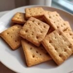 A close-up of several square, golden brown Graham Cracker Cookies stacked on a white plate.