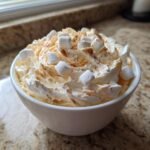 A white bowl filled with creamy Peanut Butter Fluff, topped with whipped cream, mini marshmallows, and cinnamon.