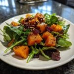 A close-up of a Roasted Butternut Squash Salad featuring bright orange cubes, mixed greens, pecans, and dried cranberries.