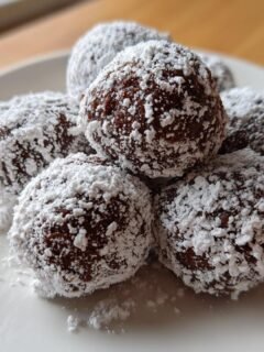 A stack of rich, dark Puppy Chow Chocolate Truffles heavily dusted with white powdered sugar on a white plate.