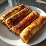 Four golden brown Sausage French Toast Roll Ups arranged diagonally on a white plate, freshly cooked.