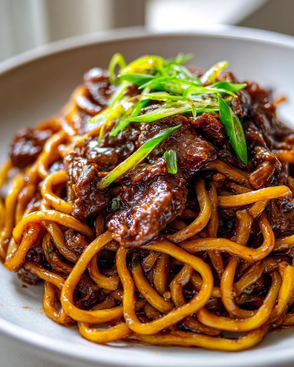 A close-up of a bowl of glossy Sticky Beef Noodles topped with tender beef pieces and fresh green onions.